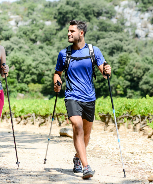 Zwei Menschen wandern mit Stöcken auf einem sonnigen Weg, umgeben von Grün und Bäumen, und lächeln sich an.