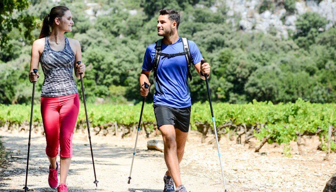 Two people are walking with sticks on a sunny path, surrounded by greenery and trees, smiling at each other.