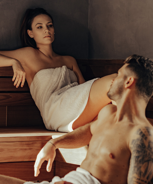 A woman in a towel and a shirtless man sit opposite each other in a sauna.